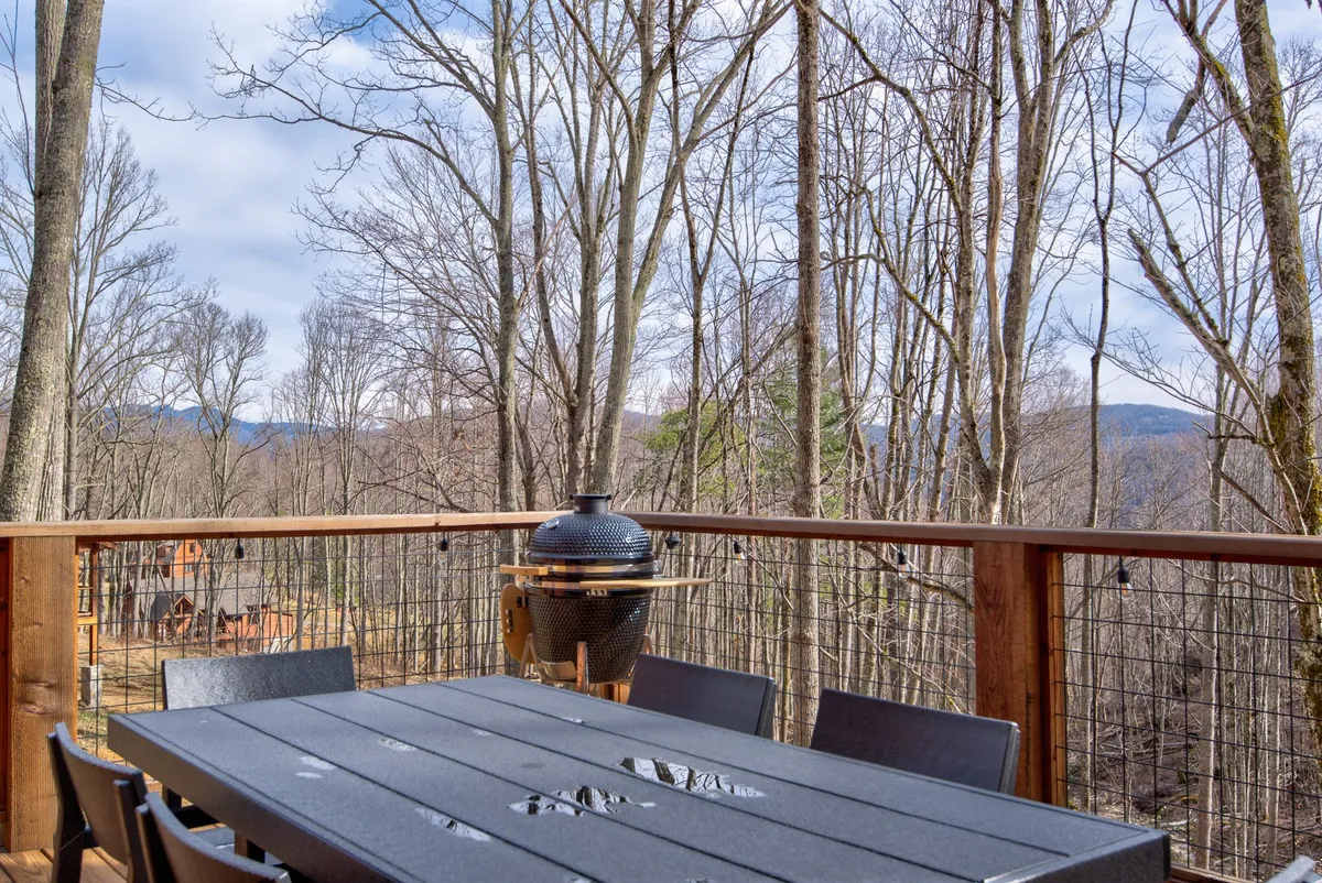 This image captures an outdoor dining table and grill nestled under a tree canopy, with a sweeping mountain view in the background. The scene is set for an alfresco meal or a celebration, surrounded by natural beauty and the inviting mountain landscape.