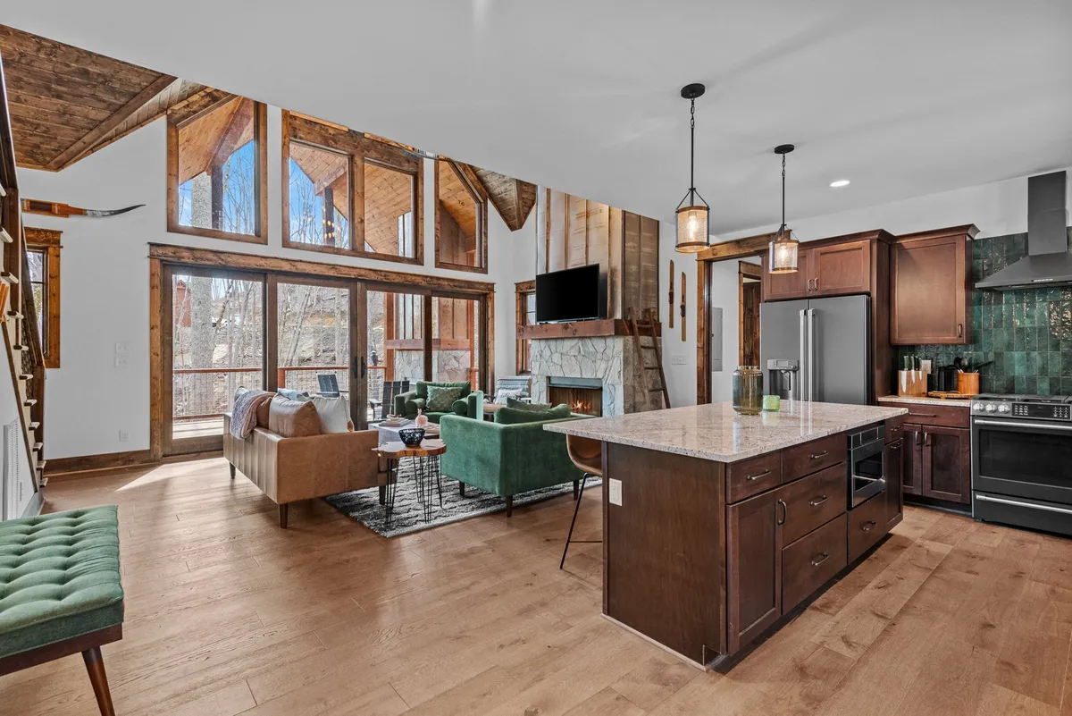 An open-concept living area featuring a stone fireplace, large windows, an island kitchen in the background, and modern, comfortable seating. The sunlight fills the space, emphasizing the room’s versatility for both relaxation and lively gatherings. The design highlights an effortless flow between living, dining, and kitchen zones.