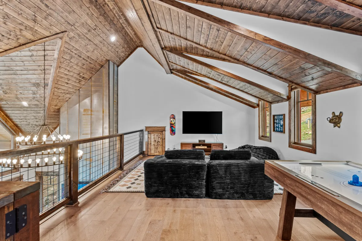 A spacious loft featuring a vaulted wooden ceiling, black plush sofas arranged for group seating, a large television for movie nights, and an air hockey table. Decorative wall art adds personality, while the relaxed layout invites both lounging and lively games.