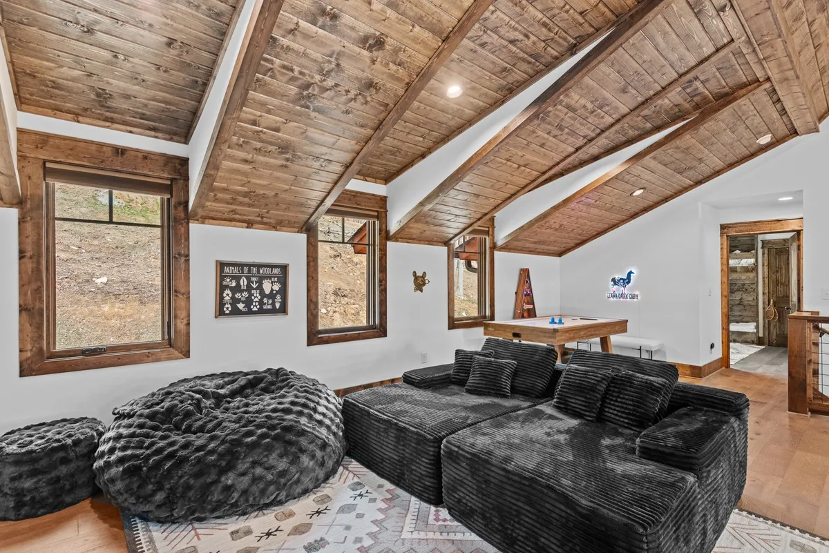 A cozy loft area with a plush black sectional sofa, bean bags for casual lounging, exposed wooden beams overhead, and large windows letting in plenty of natural light. The space feels comfortable and inviting, perfect for relaxation or group activities.