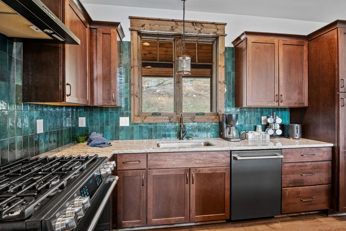 This image displays a kitchen with granite countertops, a vibrant green tiled backsplash, wooden cabinets, and a window allowing natural light to brighten the space. The scene conveys a cozy and welcoming atmosphere, suggesting a space perfect for both meal prep and casual gathering.