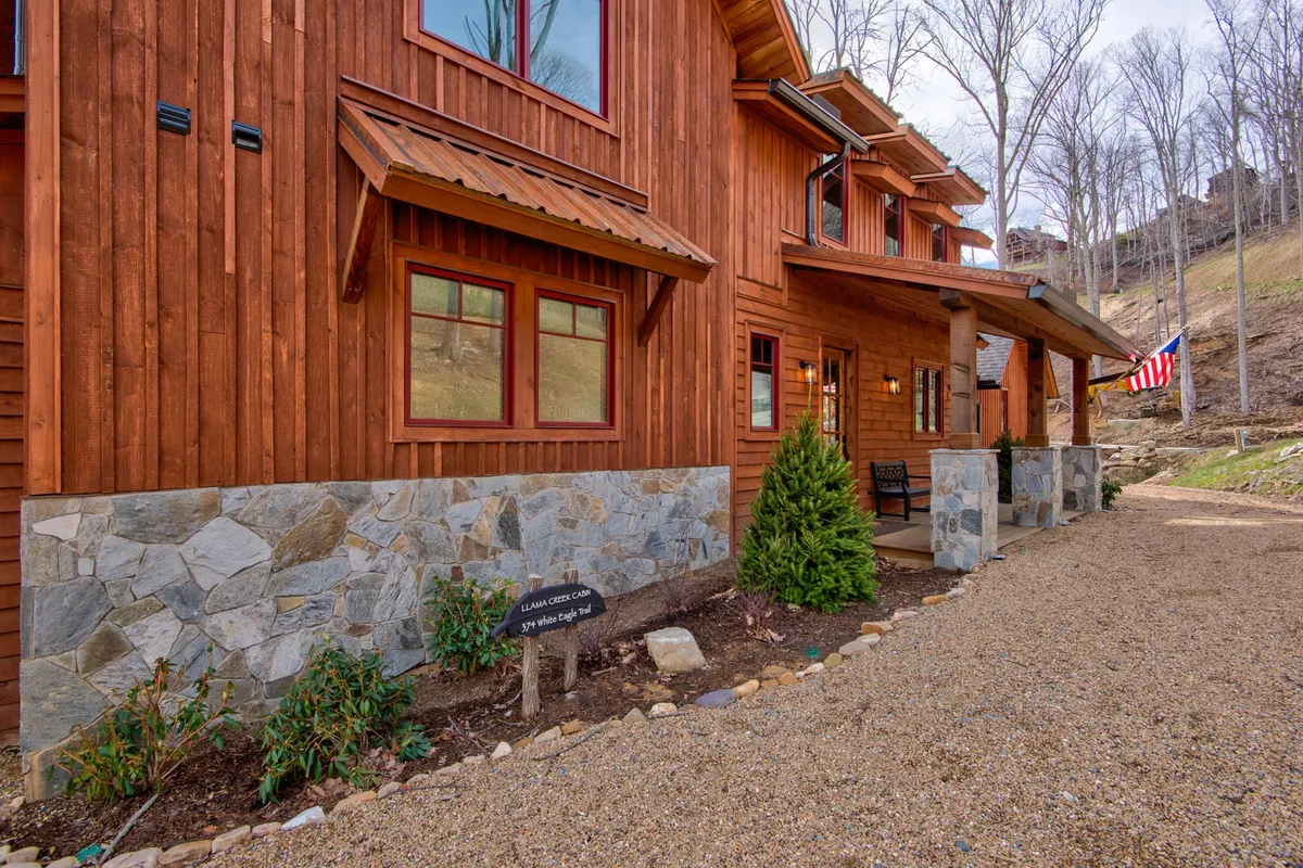 This exterior image showcases the wooden facade of a cabin with a stone foundation, a welcoming front porch, and a flag fluttering gently. The porch is set up for gatherings, with plenty of space to relax and enjoy the fresh mountain air, highlighting the spirit of togetherness and outdoor living.