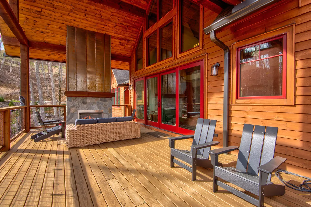This image features the exterior of a cabin with large glass windows, a stone fireplace, and a spacious wooden deck. Comfortable seating is visible, perfect for relaxing after a day of adventure. The scene conveys an inviting, cozy atmosphere, ideal for gathering family on the deck for sunset views or stargazing.