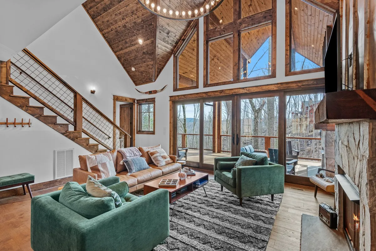 A warm and inviting interior scene with large windows framing a stunning outdoor view. The cozy seating area and modern chandelier surround a glowing fireplace, creating an atmosphere perfect for relaxing or intimate conversations while taking in the sunset.