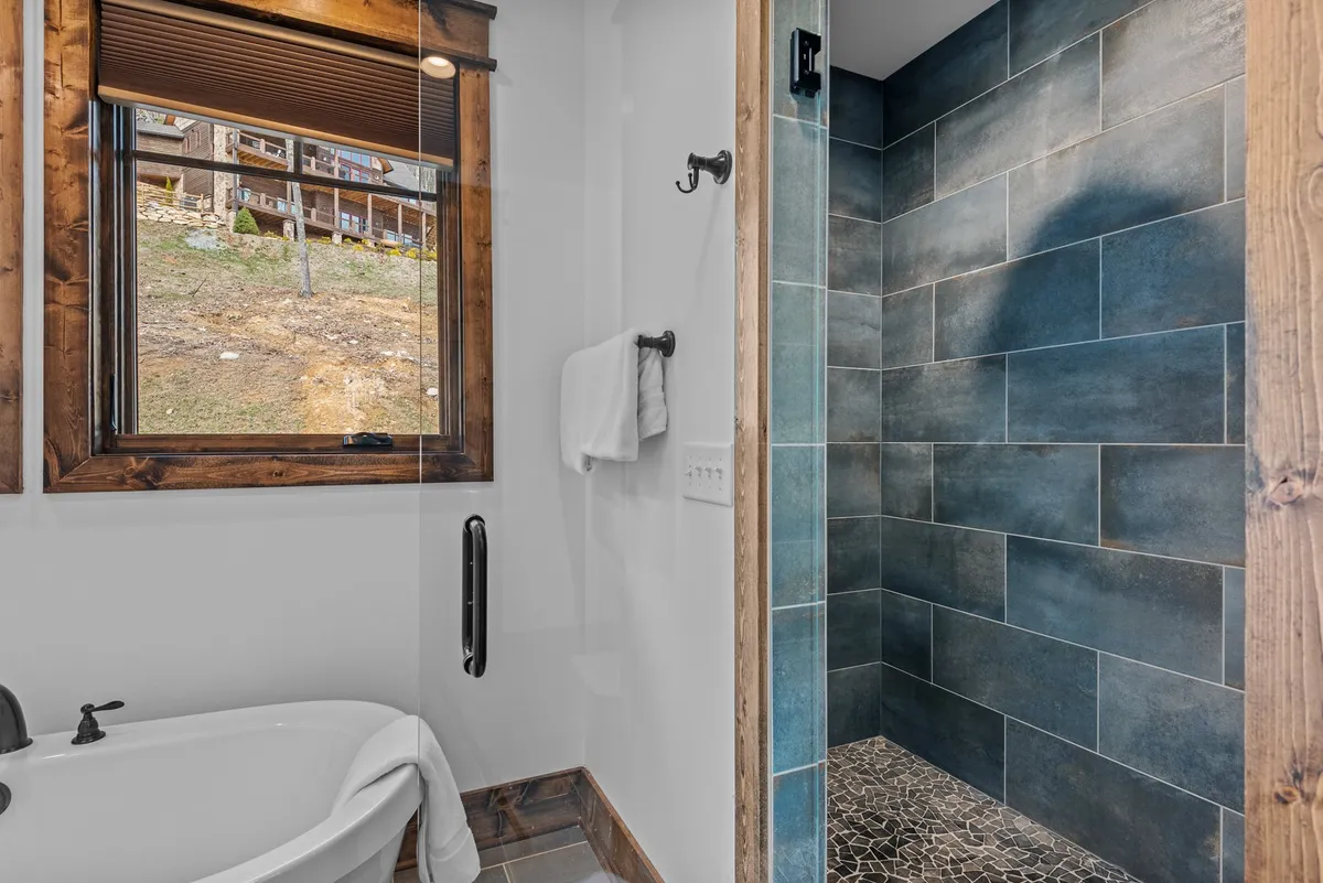 A tranquil spa-like bathroom featuring a freestanding bathtub positioned beside a large window, which allows for peaceful gazing over the surrounding nature. The shower area is finished with textured tiles, enhancing the sense of luxury and relaxation.