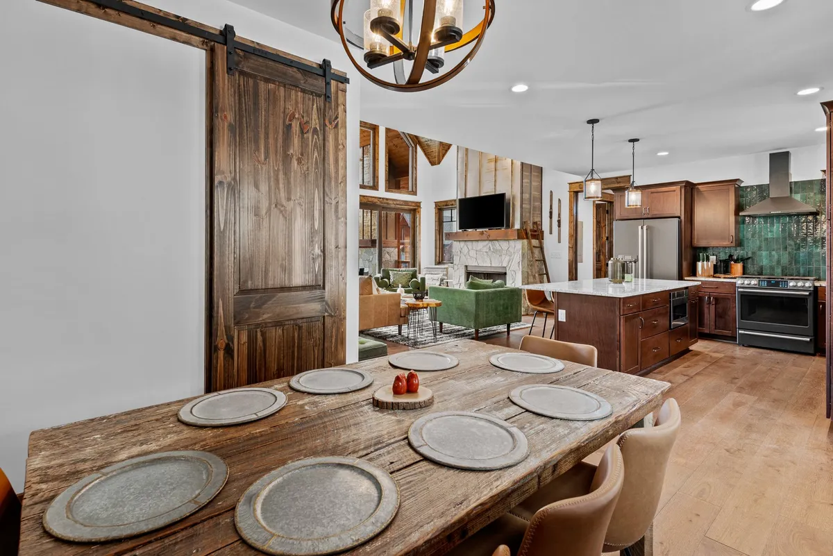 A warm, rustic dining area with a large table seating eight, a glowing chandelier overhead, and a kitchen island visible in the background. The scene hints at joyful family meals and entertaining guests in a communal, inviting setting.