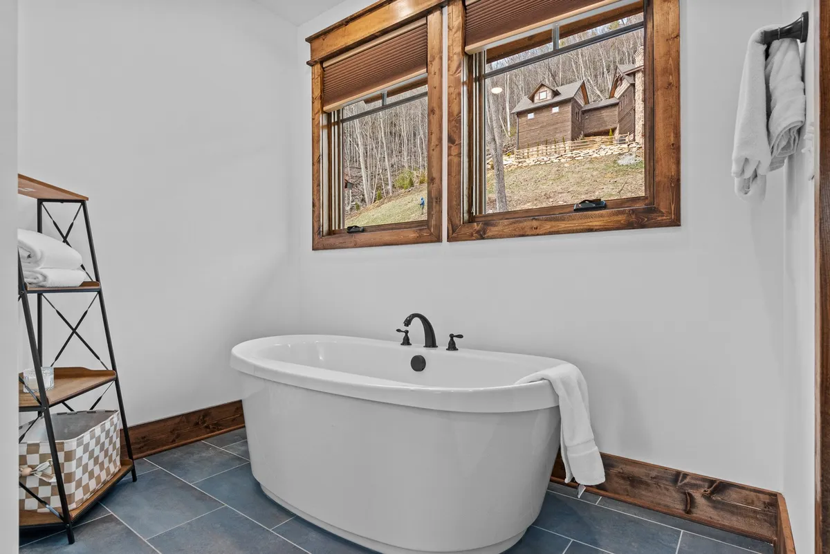 A tranquil bathroom with a freestanding soaking tub beside expansive windows, offering a peaceful view of the outdoors. Wooden accents and abundant natural light create a spa-like, inviting retreat perfect for relaxation.