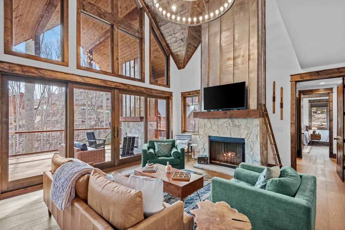 A spacious great room with soaring ceilings, large windows letting in abundant natural light, a modern chandelier, plush sofas arranged around a central coffee table, and a stone fireplace. The scene radiates warmth, comfort, and invites group gatherings. The flat-screen TV is mounted above the fireplace, blending seamlessly with the decor.