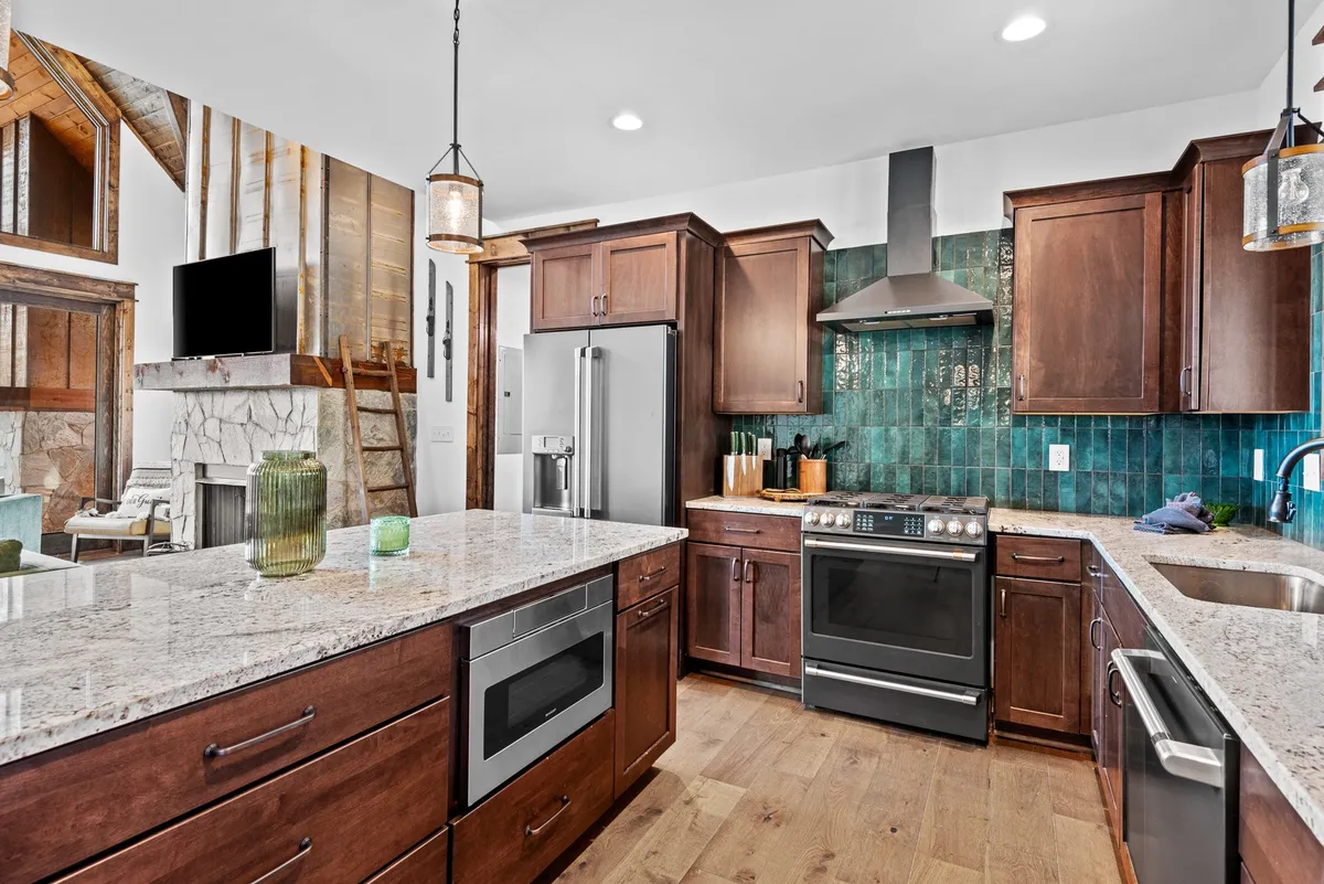 A bright, modern kitchen scene featuring granite countertops, dark wood cabinets, a green tile backsplash, and sleek stainless steel appliances. The layout is open, inviting, and perfect for gatherings or family cooking sessions.