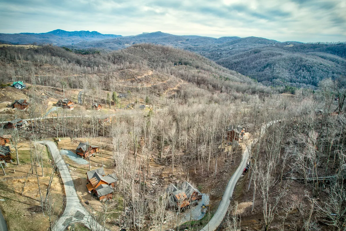 A tranquil view of rustic cabins nestled in the rolling landscape, with a winding road leading off into the distance and majestic mountains rising in the background. The natural setting is surrounded by woodland, suggesting abundant opportunities for hiking, biking, and outdoor exploration.